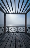 A wooden pergola structure overlooks a calm sea, with sunlight filtering through the slats creating shadows on the wooden deck. The sky is clear, and the horizon line is visible, blending into the serene ocean.