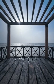 A wooden pergola structure overlooks a calm sea, with sunlight filtering through the slats creating shadows on the wooden deck. The sky is clear, and the horizon line is visible, blending into the serene ocean.
