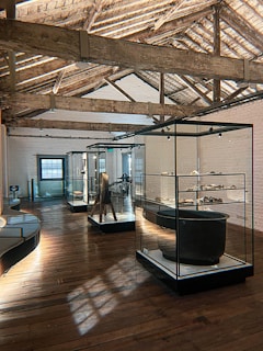 A spacious museum gallery with wooden floors and exposed wooden beams on the ceiling. Glass display cases contain various artifacts, and soft natural light filters through large windows.