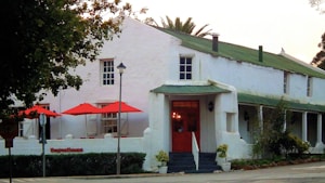 A charming, rustic building with whitewashed walls and a green roof, featuring a prominent red door at the entrance. The front of the building has red patio umbrellas, and there are small shrubs and trees nearby. A lamppost and street signs are visible in the foreground.