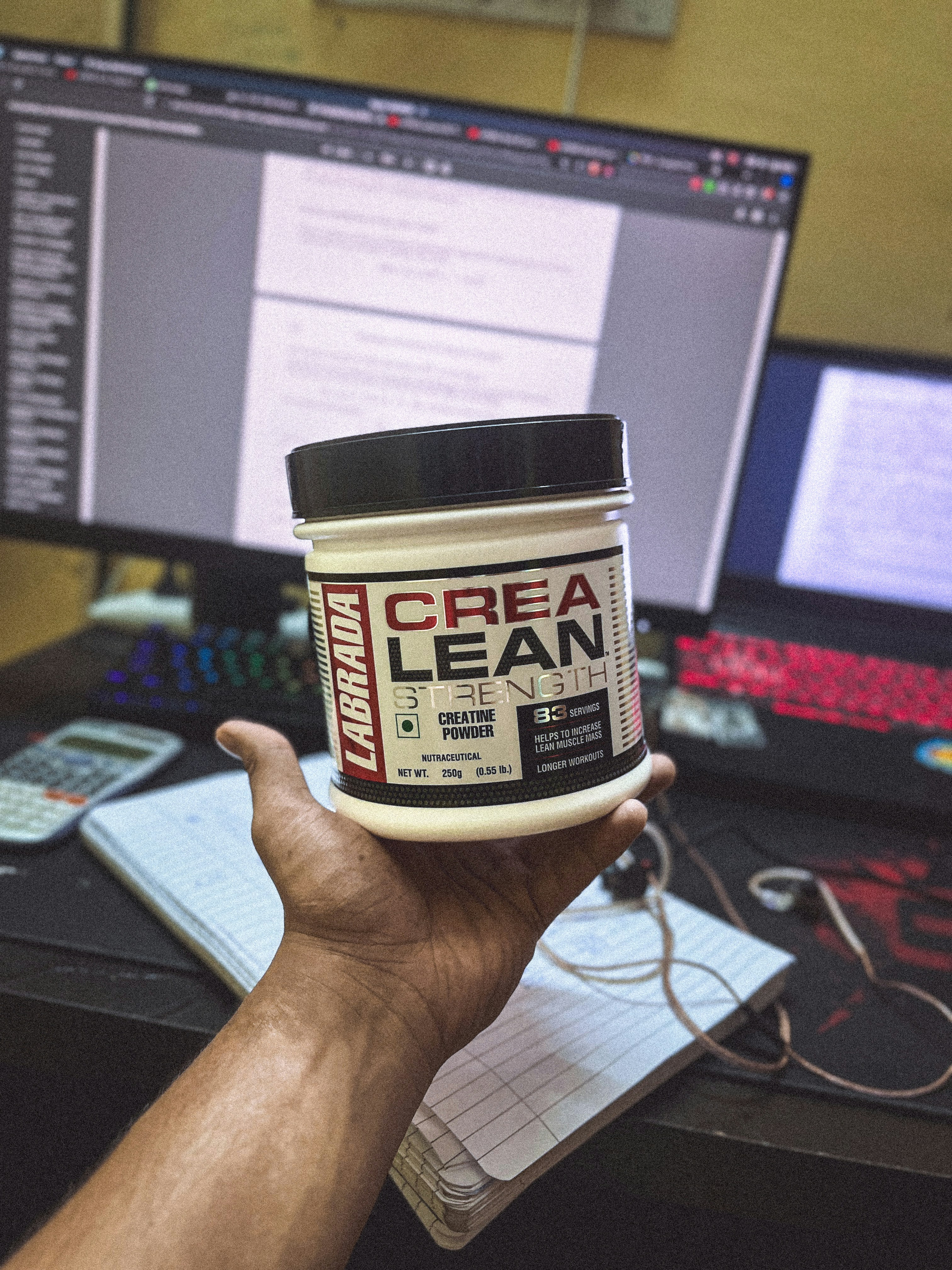 A hand holding a container of Labrada CREA LEAN strength creatine powder in front of a computer setup. The background includes a monitor displaying text, a colorful keyboard, a calculator, and a notepad with a pen.