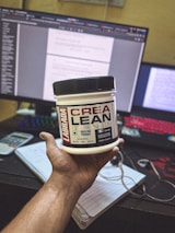 A hand holding a container of Labrada CREA LEAN strength creatine powder in front of a computer setup. The background includes a monitor displaying text, a colorful keyboard, a calculator, and a notepad with a pen.