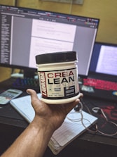 A hand holding a container of Labrada CREA LEAN strength creatine powder in front of a computer setup. The background includes a monitor displaying text, a colorful keyboard, a calculator, and a notepad with a pen.