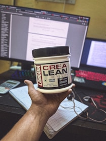 A hand holding a container of Labrada CREA LEAN strength creatine powder in front of a computer setup. The background includes a monitor displaying text, a colorful keyboard, a calculator, and a notepad with a pen.