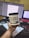 A hand holding a container of Labrada CREA LEAN strength creatine powder in front of a computer setup. The background includes a monitor displaying text, a colorful keyboard, a calculator, and a notepad with a pen.