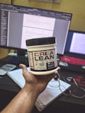 A hand holding a container of Labrada CREA LEAN strength creatine powder in front of a computer setup. The background includes a monitor displaying text, a colorful keyboard, a calculator, and a notepad with a pen.