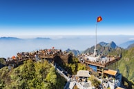 A mountain summit crowded with people enjoying a scenic viewpoint, featuring a large viewing platform adorned with a prominent red flag with a yellow star. Surrounding the platform are lush green mountains under a clear blue sky.