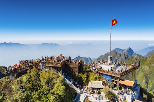 A mountain summit crowded with people enjoying a scenic viewpoint, featuring a large viewing platform adorned with a prominent red flag with a yellow star. Surrounding the platform are lush green mountains under a clear blue sky.