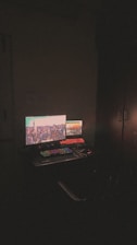 A dimly lit room with a computer setup featuring dual monitors. The main monitor displays a cityscape with bright lights, while the secondary monitor shows a software interface. A colorful RGB keyboard is visible along with a mouse and some cables on the desk.