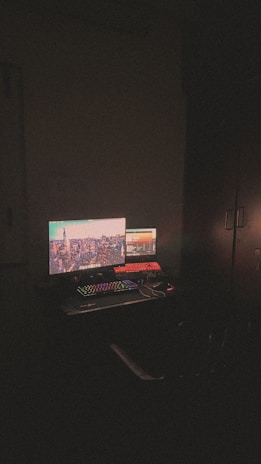 A dimly lit room with a computer setup featuring dual monitors. The main monitor displays a cityscape with bright lights, while the secondary monitor shows a software interface. A colorful RGB keyboard is visible along with a mouse and some cables on the desk.