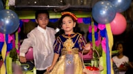 Two children dressed in traditional attire are standing in a festive setting decorated with colorful balloons and paper decorations. The girl is wearing a vibrant dress with blue and gold colors, holding a basket with red objects. The boy is dressed in a white embroidered shirt.