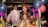Two children dressed in traditional attire are standing in a festive setting decorated with colorful balloons and paper decorations. The girl is wearing a vibrant dress with blue and gold colors, holding a basket with red objects. The boy is dressed in a white embroidered shirt.