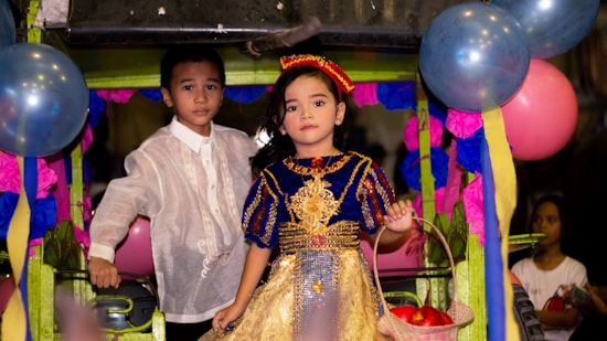 Two children dressed in traditional attire are standing in a festive setting decorated with colorful balloons and paper decorations. The girl is wearing a vibrant dress with blue and gold colors, holding a basket with red objects. The boy is dressed in a white embroidered shirt.
