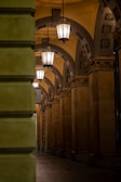 A beautifully lit architectural corridor with ornate columns and decorative arches. The hanging lanterns provide warm illumination, highlighting the intricate details of the arch design and the richly decorated capitals on the columns.