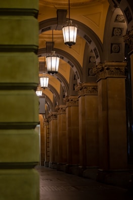 A beautifully lit architectural corridor with ornate columns and decorative arches. The hanging lanterns provide warm illumination, highlighting the intricate details of the arch design and the richly decorated capitals on the columns.