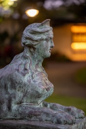 A detailed statue of a mythical creature with a human-like face, leonine body, and intricate detailing in the hair and paws. The statue appears weathered with a patina, set against an out-of-focus, warmly lit background.