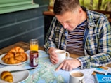 A person is studying a map spread out on a wooden table while holding a cup of coffee. The person is wearing a plaid shirt over a striped t-shirt. Nearby on the table are croissants on a white plate, a jar of jam, and a glass of orange juice. The setting appears to be outdoors with a relaxed and exploratory atmosphere.