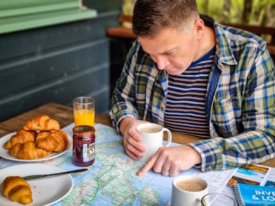 A person is studying a map spread out on a wooden table while holding a cup of coffee. The person is wearing a plaid shirt over a striped t-shirt. Nearby on the table are croissants on a white plate, a jar of jam, and a glass of orange juice. The setting appears to be outdoors with a relaxed and exploratory atmosphere.