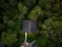 Aerial view of a house with a dark roof surrounded by dense green forest. A gravel path leads up to the house, and there is a small vehicle visible near the bottom of the image. The forest canopy covers most of the area, creating a lush, natural setting.