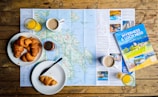 On a wooden table lies a detailed map surrounded by breakfast items. Freshly baked croissants are placed on white plates, accompanied by cups of coffee and glasses of orange juice. A travel guidebook titled 'Inverness & Loch Ness' rests on the map, suggesting a planned trip to the region.