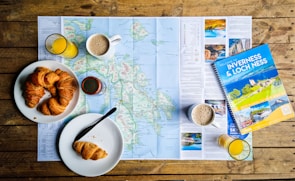 On a wooden table lies a detailed map surrounded by breakfast items. Freshly baked croissants are placed on white plates, accompanied by cups of coffee and glasses of orange juice. A travel guidebook titled 'Inverness & Loch Ness' rests on the map, suggesting a planned trip to the region.