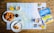 On a wooden table lies a detailed map surrounded by breakfast items. Freshly baked croissants are placed on white plates, accompanied by cups of coffee and glasses of orange juice. A travel guidebook titled 'Inverness & Loch Ness' rests on the map, suggesting a planned trip to the region.