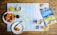 On a wooden table lies a detailed map surrounded by breakfast items. Freshly baked croissants are placed on white plates, accompanied by cups of coffee and glasses of orange juice. A travel guidebook titled 'Inverness & Loch Ness' rests on the map, suggesting a planned trip to the region.