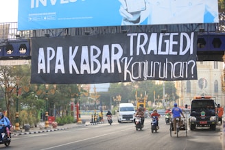 A large black banner with white text hangs over a busy street. Below, several motorcyclists and a person on a pedicab move along the road, accompanied by a black truck. The street is lined with decorative street lamps and some greenery.