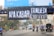 A large black banner with white text hangs over a busy street. Below, several motorcyclists and a person on a pedicab move along the road, accompanied by a black truck. The street is lined with decorative street lamps and some greenery.