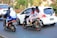 Two motorcycles are on a busy street, each with two individuals. The motorcycle on the left has two people wearing a helmet, with the passenger in a blue and white uniform. The motorcycle on the right has two individuals wearing similar outfits with hijabs. There are cars nearby and lush green vegetation in the background, along with Indonesian flags.