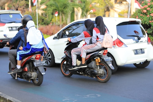 Two motorcycles are on a busy street, each with two individuals. The motorcycle on the left has two people wearing a helmet, with the passenger in a blue and white uniform. The motorcycle on the right has two individuals wearing similar outfits with hijabs. There are cars nearby and lush green vegetation in the background, along with Indonesian flags.