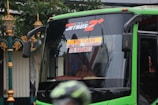 A green bus with signage indicating its route from Malang to Surabaya via Tol Karanglo. The front of the bus is visible, with reflections on the windshield. A person is seated inside the bus with part of their hand visible through a small window.