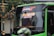 A green bus with signage indicating its route from Malang to Surabaya via Tol Karanglo. The front of the bus is visible, with reflections on the windshield. A person is seated inside the bus with part of their hand visible through a small window.
