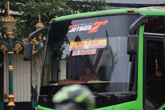 A green bus with signage indicating its route from Malang to Surabaya via Tol Karanglo. The front of the bus is visible, with reflections on the windshield. A person is seated inside the bus with part of their hand visible through a small window.
