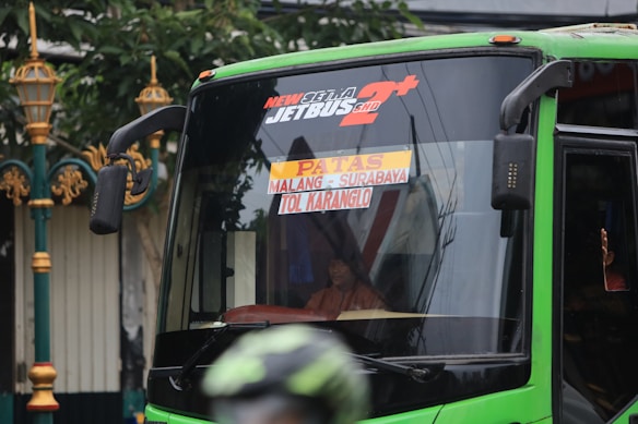 A green bus with signage indicating its route from Malang to Surabaya via Tol Karanglo. The front of the bus is visible, with reflections on the windshield. A person is seated inside the bus with part of their hand visible through a small window.
