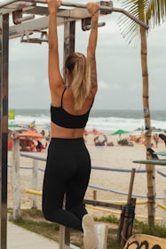 A person with tattoos is seen from behind, performing a pull-up on a metal bar at an outdoor gym located on a beach. They are wearing a black sports bra and leggings while their hair is tied in a ponytail. Below, there are beachgoers sitting under colorful umbrellas near the ocean.