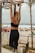 A person with tattoos is seen from behind, performing a pull-up on a metal bar at an outdoor gym located on a beach. They are wearing a black sports bra and leggings while their hair is tied in a ponytail. Below, there are beachgoers sitting under colorful umbrellas near the ocean.