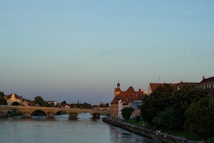 A picturesque riverside scene featuring a historic stone bridge stretching across the calm water. On the right, traditional European buildings with red-tiled roofs are bordered by lush green trees. The serene sky is transitioning from late afternoon light, casting a warm glow over the landscape.