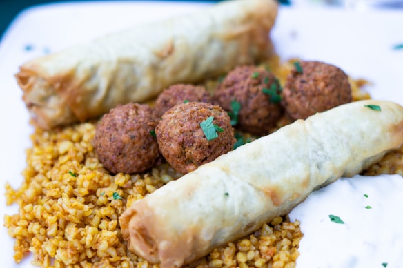 A dish featuring golden cooked bulgur spread across the plate, accompanied by crispy fried croquettes garnished with chopped herbs. Two flaky pastry rolls are placed on top, with a dollop of yogurt sauce on the side.