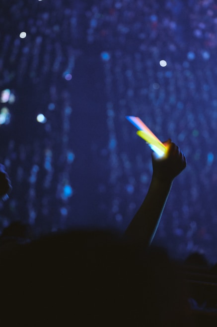 Hands holding a Korean lightstick illuminating a concert crowd under colorful stage lights