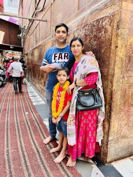 A family of three stands in a narrow, bustling alley. The man wears a blue shirt and jeans, holding papers, while the woman is dressed in a pink traditional outfit with a patterned shawl and holding a black handbag. Their young child is in a red shirt and jeans, adorned with a marigold garland. The wall behind them is textured and brown, and there are people and a scooter in the background.