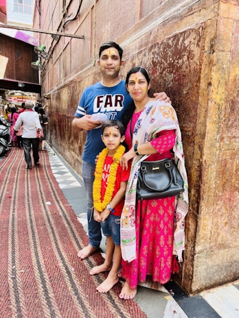 A family of three stands in a narrow, bustling alley. The man wears a blue shirt and jeans, holding papers, while the woman is dressed in a pink traditional outfit with a patterned shawl and holding a black handbag. Their young child is in a red shirt and jeans, adorned with a marigold garland. The wall behind them is textured and brown, and there are people and a scooter in the background.