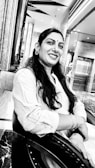 A woman with long hair and a bindi is smiling while sitting on a chair. She is wearing a light-colored traditional outfit and earrings. The background features a modern interior with marble surfaces and a mirrored wall.