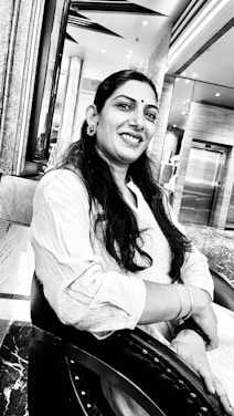 A woman with long hair and a bindi is smiling while sitting on a chair. She is wearing a light-colored traditional outfit and earrings. The background features a modern interior with marble surfaces and a mirrored wall.