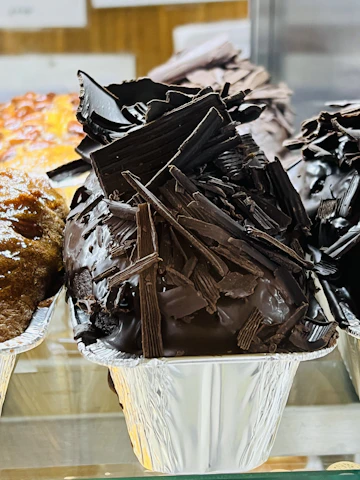 A sumptuous display of a chocolate dessert covered with a cascade of dark chocolate shavings. The dessert is placed in a shiny foil container, highlighting the rich texture of the chocolate. Behind it, there is another dessert with a caramelized top, offering a contrast in color and texture.
