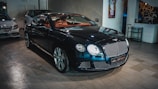 A sleek, luxurious black car with shiny chrome details is parked indoors. The vehicle features a prominent front grille and distinctive headlights. There are other cars present in the background, suggesting a showroom or garage setting. The tiled floor and wall art add to the upscale ambiance.