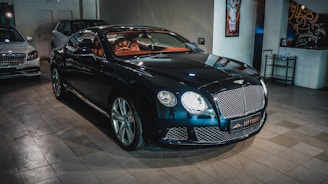 A sleek, luxurious black car with shiny chrome details is parked indoors. The vehicle features a prominent front grille and distinctive headlights. There are other cars present in the background, suggesting a showroom or garage setting. The tiled floor and wall art add to the upscale ambiance.