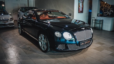A sleek, luxurious black car with shiny chrome details is parked indoors. The vehicle features a prominent front grille and distinctive headlights. There are other cars present in the background, suggesting a showroom or garage setting. The tiled floor and wall art add to the upscale ambiance.