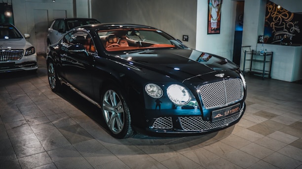 A sleek, luxurious black car with shiny chrome details is parked indoors. The vehicle features a prominent front grille and distinctive headlights. There are other cars present in the background, suggesting a showroom or garage setting. The tiled floor and wall art add to the upscale ambiance.