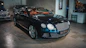 A sleek, luxurious black car with shiny chrome details is parked indoors. The vehicle features a prominent front grille and distinctive headlights. There are other cars present in the background, suggesting a showroom or garage setting. The tiled floor and wall art add to the upscale ambiance.